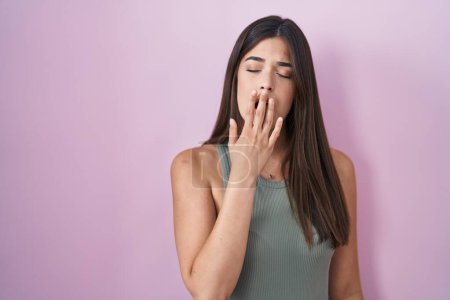 Foto de Mujer hispana de pie sobre fondo rosa aburrido bostezar cansado cubriendo la boca con la mano. inquietud y somnolencia. - Imagen libre de derechos