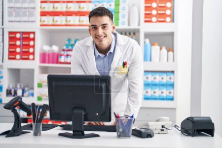 Foto de Joven farmacéutico hispano sonriendo confiado usando computadora en farmacia - Imagen libre de derechos