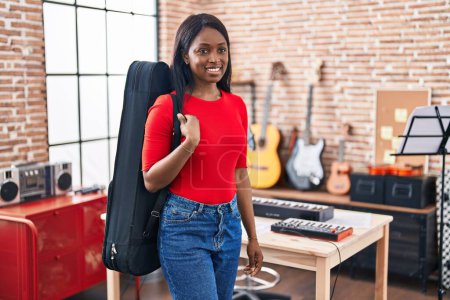 Photo for African young woman wearing guitar case at music studio looking positive and happy standing and smiling with a confident smile showing teeth - Royalty Free Image