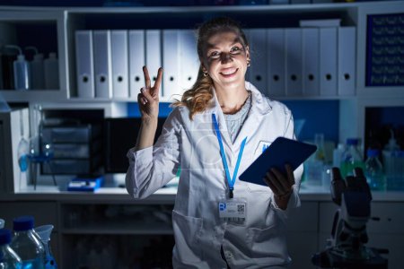 Téléchargez les photos : Beautiful blonde woman working at scientist laboratory late at night smiling with happy face winking at the camera doing victory sign with fingers. number two. - en image libre de droit