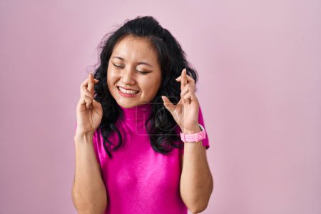 Foto de Mujer asiática joven de pie sobre fondo rosa gesto dedo cruzado sonriendo con esperanza y los ojos cerrados. suerte y concepto supersticioso. - Imagen libre de derechos