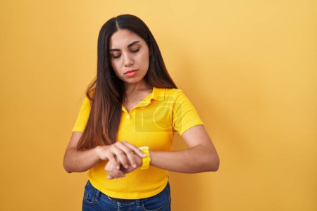 Foto de Mujer árabe joven de pie sobre fondo amarillo comprobar la hora en el reloj de pulsera, relajado y seguro - Imagen libre de derechos