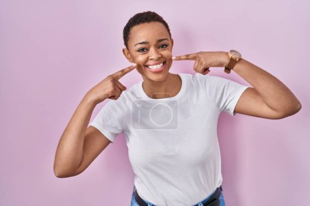 Téléchargez les photos : Belle femme afro-américaine debout sur fond rose souriant et joyeux montrant et pointant avec les doigts les dents et la bouche. concept de santé dentaire. - en image libre de droit