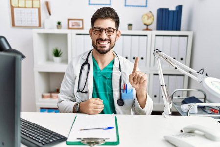 Photo for Young man with beard wearing doctor uniform and stethoscope at the clinic showing and pointing up with finger number one while smiling confident and happy. - Royalty Free Image
