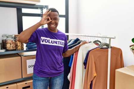 Foto de Joven africano vistiendo camiseta voluntaria en las donaciones de pie sonriendo feliz haciendo signo bien con la mano en el ojo mirando a través de los dedos - Imagen libre de derechos