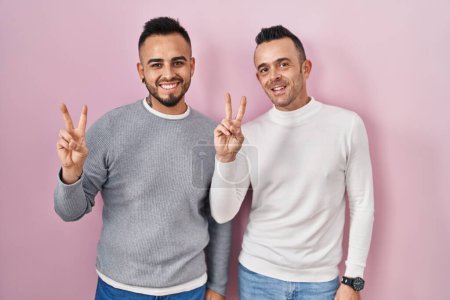 Téléchargez les photos : Couple homosexuel debout sur fond rose souriant avec le visage heureux clin d'oeil à la caméra faisant signe de victoire avec les doigts. numéro deux. - en image libre de droit
