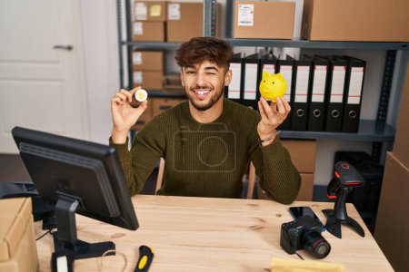 Téléchargez les photos : Homme arabe avec barbe travaillant à la petite entreprise ecommerce tenant Bitcoin clin d'oeil en regardant l'appareil photo avec expression sexy, gai et heureux visage. - en image libre de droit