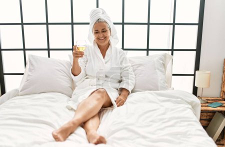 Téléchargez les photos : Middle age grey-haired woman drinking champagne sitting on the bed at bedroom. - en image libre de droit