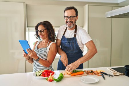 Téléchargez les photos : Moyen âge couple hispanique souriant cuisine confiante et en utilisant touchpad à la cuisine - en image libre de droit