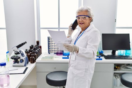 Foto de Mujer mayor de cabello gris con uniforme científico hablando en el teléfono inteligente en el laboratorio - Imagen libre de derechos