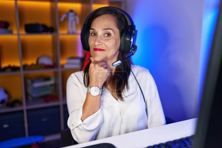 Foto de Middle age brunette woman playing video games smiling looking confident at the camera with crossed arms and hand on chin. thinking positive. - Imagen libre de derechos