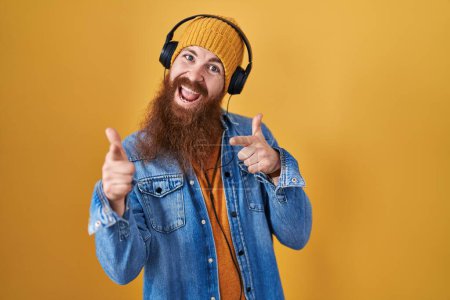 Foto de Hombre caucásico con barba larga escuchando música usando auriculares apuntando con los dedos a la cámara con cara alegre y divertida. buena energía y vibraciones. - Imagen libre de derechos