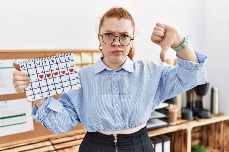 Téléchargez les photos : Young redhead woman holding heart calendar at the office with angry face, negative sign showing dislike with thumbs down, rejection concept - en image libre de droit