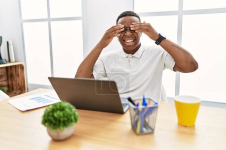 Foto de Hombre africano joven que trabaja en la oficina usando computadora portátil que cubre los ojos con las manos sonriendo alegre y divertido. concepto ciego. - Imagen libre de derechos
