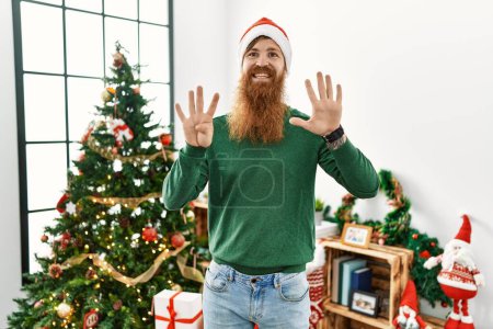 Téléchargez les photos : Homme rousse à longue barbe portant un chapeau de Noël par arbre de Noël montrant et pointant vers le haut avec les doigts numéro neuf tout en souriant confiant et heureux. - en image libre de droit