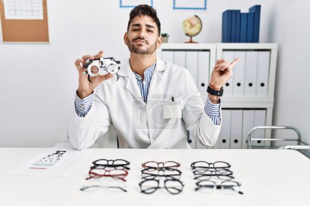 Photo for Young optician man holding optometry glasses pointing with hand finger to the side showing advertisement, serious and calm face - Royalty Free Image