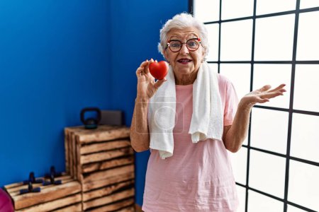 Téléchargez les photos : Femme âgée avec les cheveux gris portant des vêtements de sport tenant coeur rouge célébrant la réalisation avec sourire heureux et expression gagnante avec la main levée - en image libre de droit