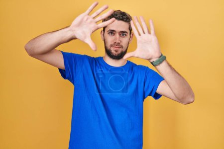 Foto de Hombre hispano con barba de pie sobre fondo amarillo haciendo marco usando manos palmas y dedos, perspectiva de la cámara - Imagen libre de derechos