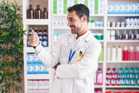 Téléchargez les photos : Bel homme hispanique travaillant à la pharmacie pharmacie à l'air fier, souriant faire pouce vers le haut geste sur le côté - en image libre de droit