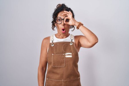 Foto de Middle age woman wearing apron over white background doing ok gesture shocked with surprised face, eye looking through fingers. unbelieving expression. - Imagen libre de derechos