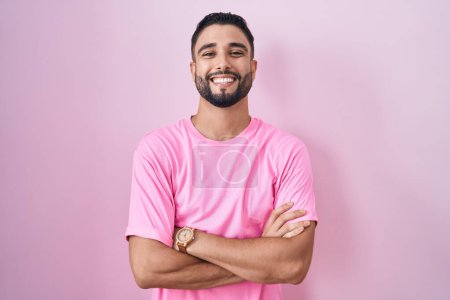 Foto de Joven hispano de pie sobre fondo rosa rostro feliz sonriendo con los brazos cruzados mirando a la cámara. persona positiva. - Imagen libre de derechos