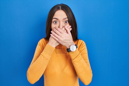 Photo for Young brunette woman standing over blue background shocked covering mouth with hands for mistake. secret concept. - Royalty Free Image