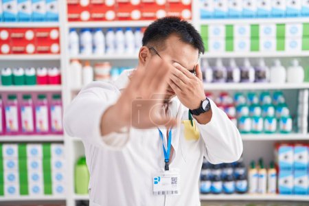 Photo for Chinese young man working at pharmacy drugstore covering eyes with hands and doing stop gesture with sad and fear expression. embarrassed and negative concept. - Royalty Free Image