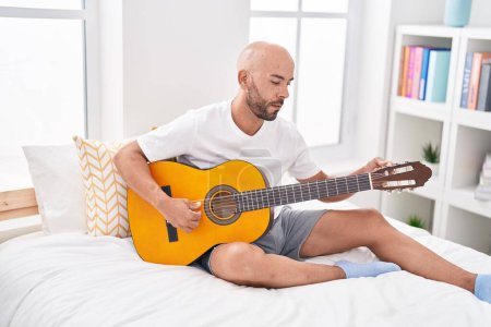 Foto de Young bald man playing classical guitar sitting on bed at bedroom - Imagen libre de derechos