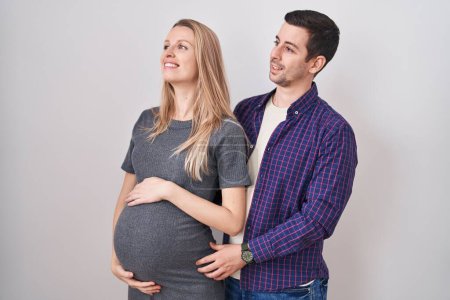 Téléchargez les photos : Jeune couple s'attendant à un bébé debout sur fond blanc regardant de côté avec le sourire sur le visage, expression naturelle. riant confiant. - en image libre de droit
