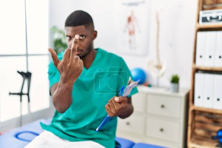 Foto de Young african american man working at pain recovery clinic showing middle finger, impolite and rude fuck off expression - Imagen libre de derechos