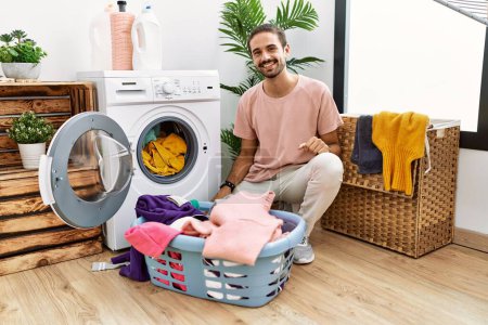 Foto de Young hispanic man putting dirty laundry into washing machine looking positive and happy standing and smiling with a confident smile showing teeth - Imagen libre de derechos