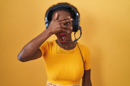 Foto de Mujer africana con el pelo rizado de pie sobre el fondo amarillo con auriculares asomándose en shock cubriendo la cara y los ojos con la mano, mirando a través de los dedos con expresión avergonzada. - Imagen libre de derechos