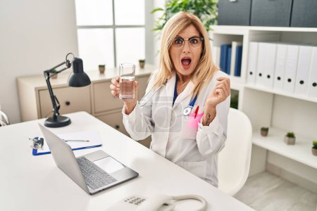 Foto de Middle age blonde doctor woman working using computer laptop drinking water scared and amazed with open mouth for surprise, disbelief face - Imagen libre de derechos