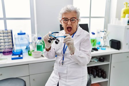 Foto de Mujer mayor con el pelo gris trabajando en el laboratorio científico usando lupas enojadas y locas gritando frustradas y furiosas, gritando con ira. rabia y concepto agresivo. - Imagen libre de derechos
