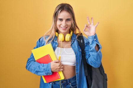 Téléchargez les photos : Young blonde woman wearing student backpack and holding books smiling positive doing ok sign with hand and fingers. successful expression. - en image libre de droit
