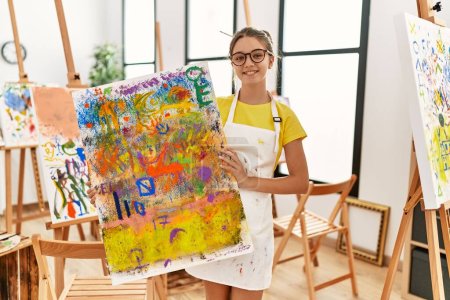 Foto de Adorable girl smiling confident holding canvas draw at art studio - Imagen libre de derechos