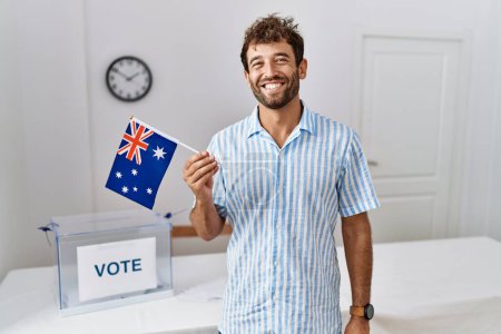Photo for Young handsome man at political campaign election holding australia flag looking positive and happy standing and smiling with a confident smile showing teeth - Royalty Free Image