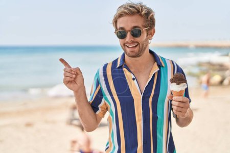 Photo for Caucasian man eating an ice cream at the beach smiling happy pointing with hand and finger to the side - Royalty Free Image