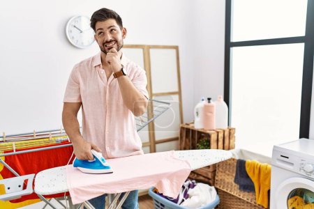 Foto de Young man with beard ironing clothes at home with hand on chin thinking about question, pensive expression. smiling and thoughtful face. doubt concept. - Imagen libre de derechos