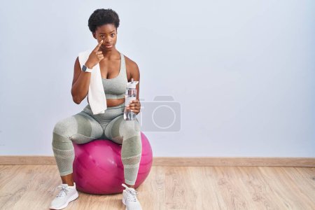 Foto de Mujer afroamericana usando ropa deportiva sentada en una pelota de pilates apuntando al ojo observándote gesto, expresión sospechosa - Imagen libre de derechos
