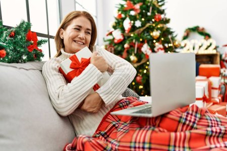 Téléchargez les photos : Moyen âge femme caucasienne en utilisant un ordinateur portable tenant cadeau assis par arbre de Noël à la maison - en image libre de droit
