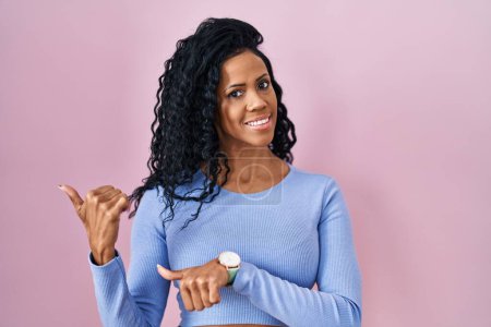 Foto de Middle age hispanic woman standing over pink background pointing to the back behind with hand and thumbs up, smiling confident - Imagen libre de derechos