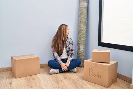 Foto de Young blonde woman sitting on the floor moving to a new home looking to side, relax profile pose with natural face with confident smile. - Imagen libre de derechos