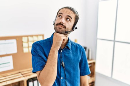 Photo for Young hispanic man with beard wearing call center agent headset at the office serious face thinking about question with hand on chin, thoughtful about confusing idea - Royalty Free Image