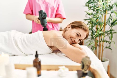Téléchargez les photos : Moyen âge femme caucasienne ayant massage du dos à l'aide d'un pistolet à percussion au centre de beauté - en image libre de droit