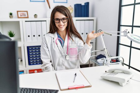 Photo for Young doctor woman wearing doctor uniform and stethoscope at the clinic smiling cheerful presenting and pointing with palm of hand looking at the camera. - Royalty Free Image