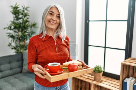 Foto de Mujer de pelo gris de mediana edad sonriendo confiada sosteniendo bandeja de desayuno en casa - Imagen libre de derechos