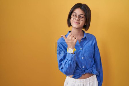 Photo for Young girl standing over yellow background smiling with happy face looking and pointing to the side with thumb up. - Royalty Free Image