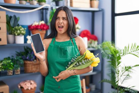 Photo for Brunette woman working at florist shop holding smartphone angry and mad screaming frustrated and furious, shouting with anger. rage and aggressive concept. - Royalty Free Image