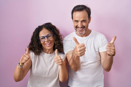 Téléchargez les photos : Couple hispanique du moyen âge ensemble sur fond rose signe de succès faisant geste positif avec la main, pouces levés souriant et heureux. expression joyeuse et geste gagnant. - en image libre de droit